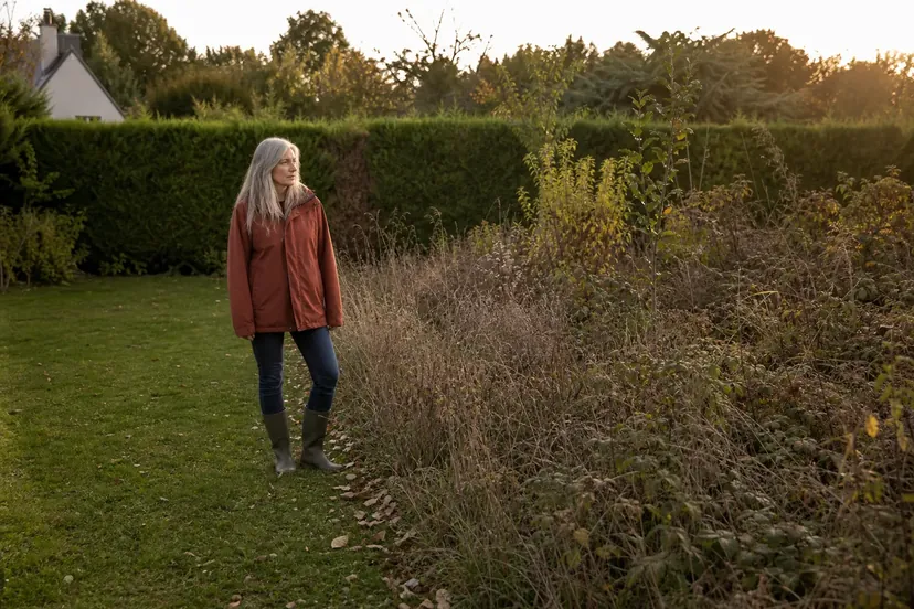 Femme aux longs cheveux gris argentés, en veste de pluie rouille et bottes de caoutchouc, debout au bord d'une pelouse qui s'arrête devant une large bande de friche aux herbes hautes sèches et aux ronces, avec une haie et une maison au fond, en fin de journée d'automne.