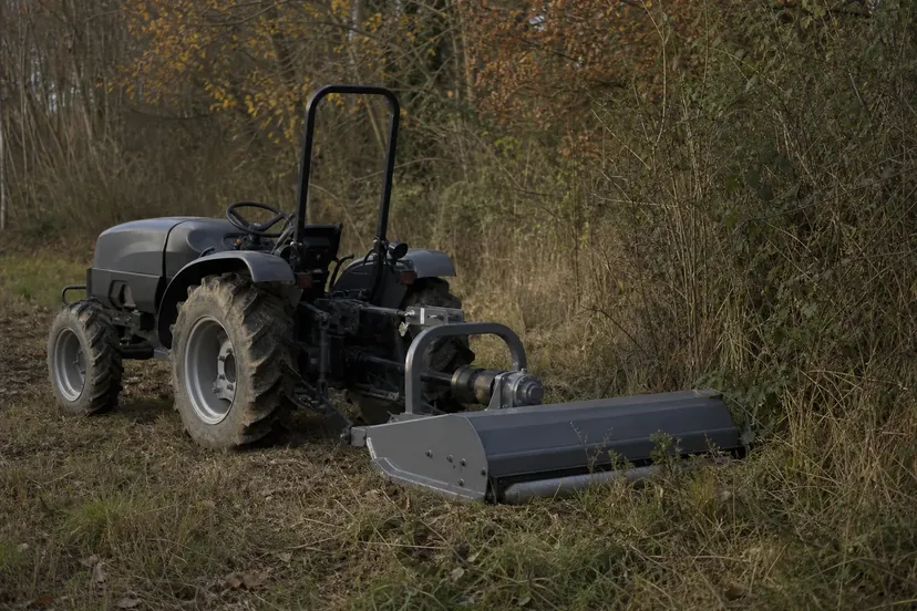 Petit tracteur gris à l'arrêt sur une parcelle d'herbes hautes sèches en lisière de bois, un broyeur attelé à l'arrière et posé au sol.