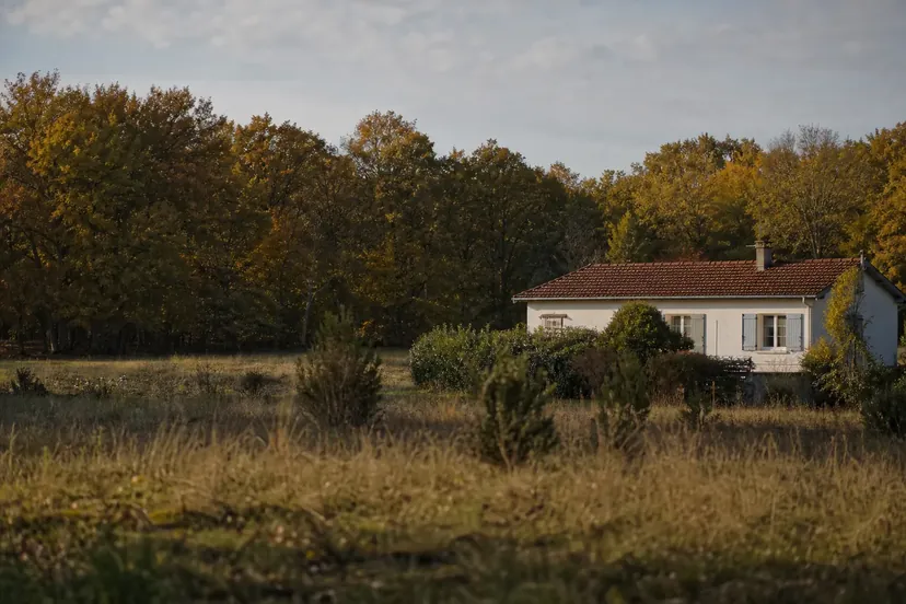 Maison de plain-pied aux volets clairs devant la lisière d'un bois de feuillus aux couleurs d'automne, séparée du bois par un pré d'herbe sèche parsemé de buissons.