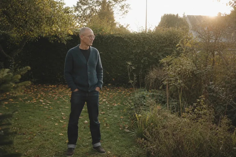 Homme au crâne rasé et aux lunettes rondes, en cardigan bleu canard, debout les mains dans les poches sur une pelouse parsemée de feuilles mortes, qui regarde des massifs envahis d'herbes hautes devant une haie, dans une lumière de fin de journée.