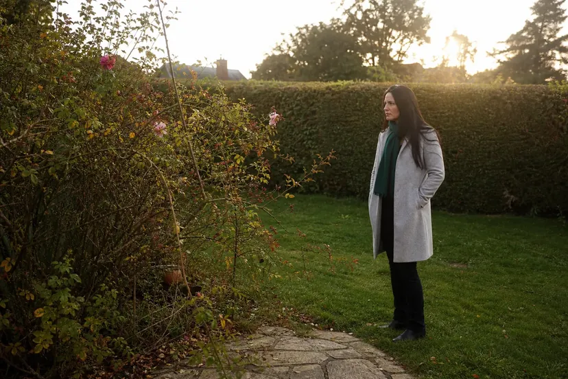 Femme aux longs cheveux noirs, en manteau gris clair et écharpe verte, debout sur une pelouse, les mains dans les poches, qui regarde un grand rosier touffu aux dernières fleurs roses et aux feuilles jaunissantes, devant une haie, en fin de journée d'automne.