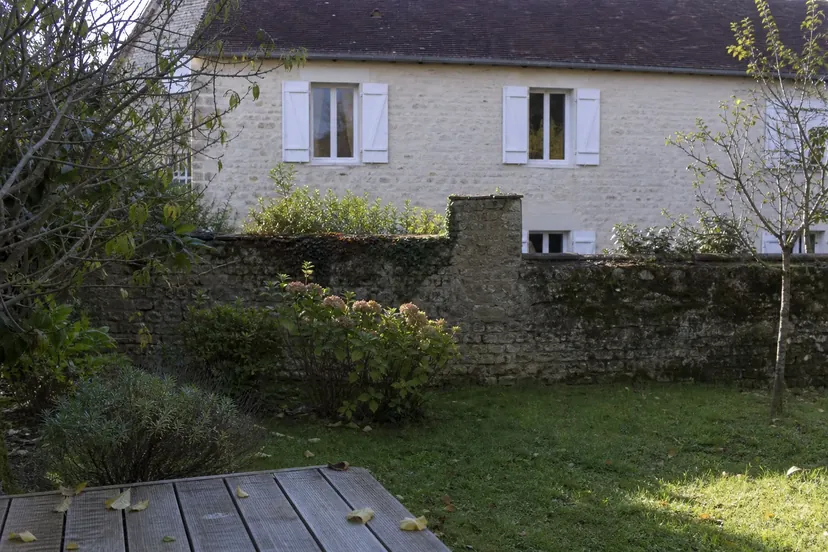 Bord d'une terrasse en bois grisé couverte de quelques feuilles mortes, devant une pelouse, un vieux mur de pierre et, au-delà, la façade en pierre claire d'une maison ancienne aux volets blancs et au toit de tuiles.