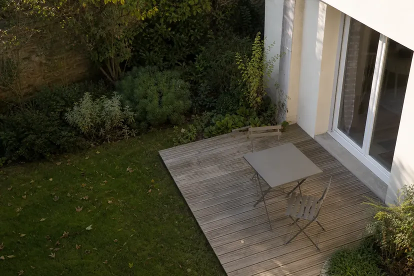 Petite terrasse en bois grisé de plain-pied contre la façade d'une maison, avec une table et deux chaises pliantes, bordée par une pelouse semée de feuilles mortes et par un massif d'arbustes, vue depuis l'étage.